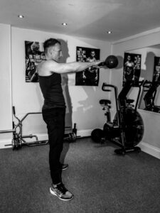 A male personal trainer in black workout clothes lifts a kettlebell in a gym.