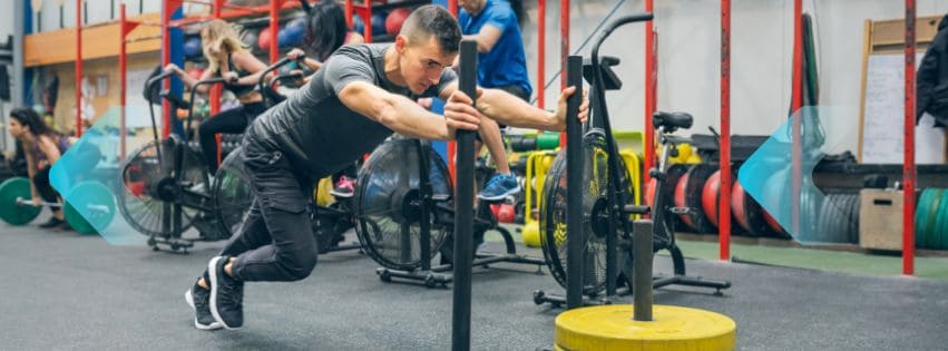 Man pushing weighted sled in a busy indoor fitness class.